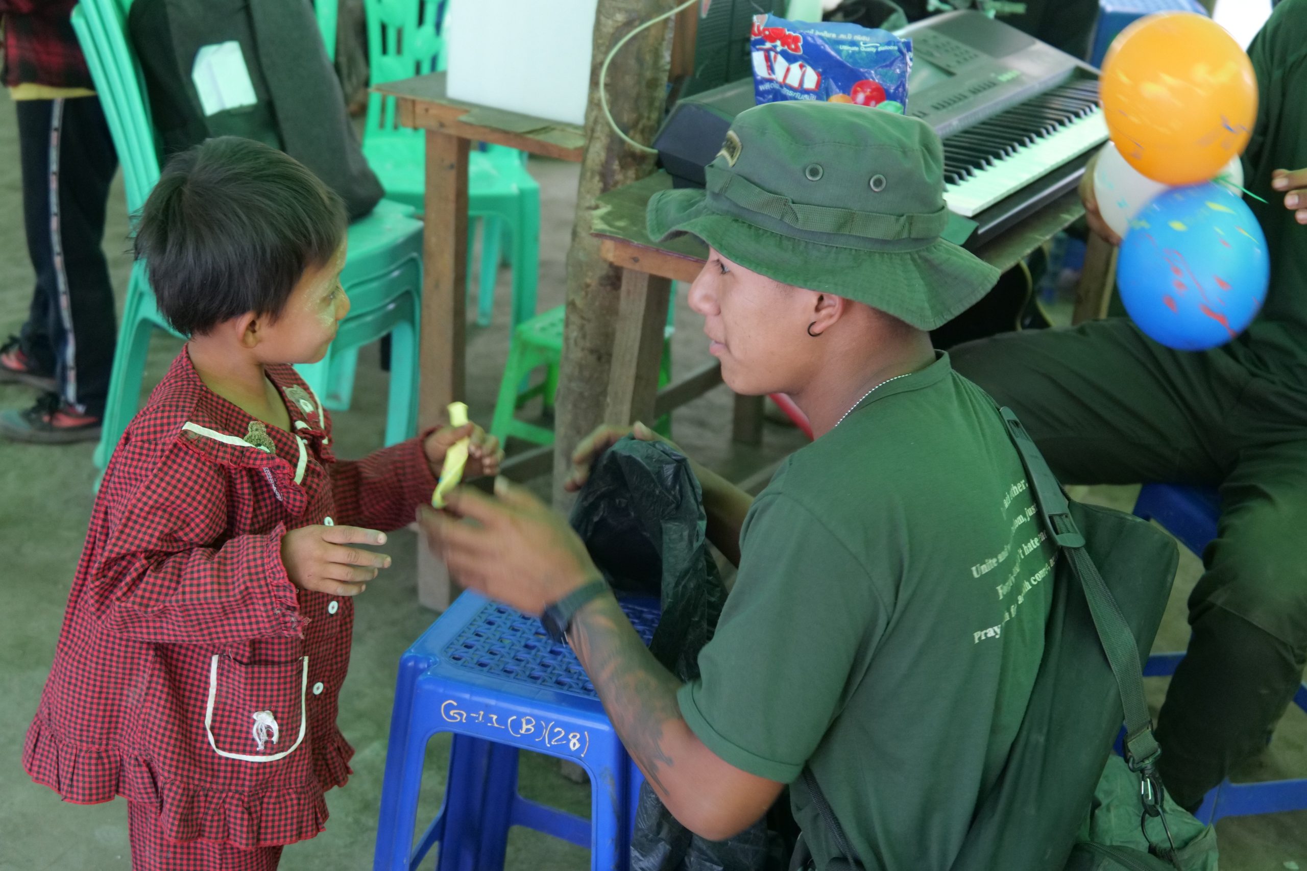 A child in a red checkered outfit interacts joyfully with a young man in a green shirt and hat, surrounded by colorful balloons and chairs.