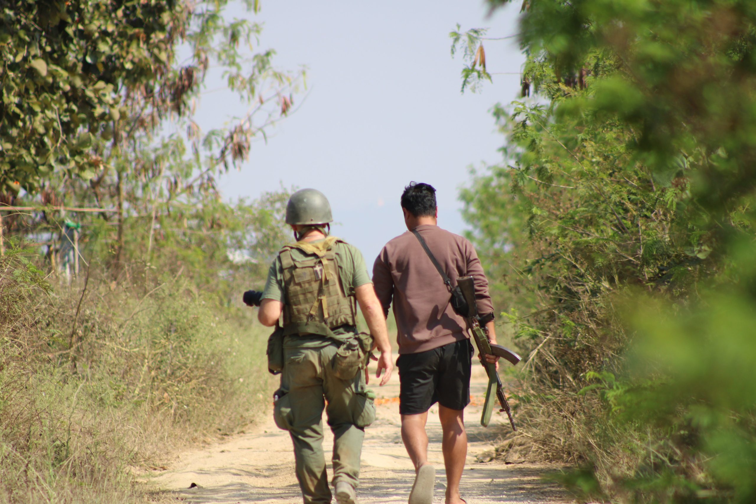 Soldier and civilian walking on a dirt path surrounded by greenery, with the soldier carrying a camera and the civilian holding a firearm.