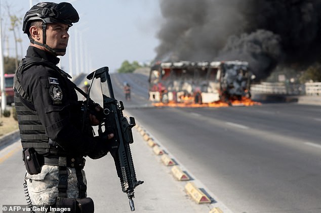 TOPSHOT - A member of the Prosecutor's Office stands guard near a burning bus at one of the main avenues after it was set on fire by organised crime groups in response to an operation in Jalisco to arrest a high-priority security target in Zapopan, state of Jalisco, Mexico, on February 22, 2026. Armed civilians blocked several roads in the state of Jalisco, in western Mexico, following an operation by federal forces in the town of Tapalpa, local authorities reported. Jalisco, which will host four matches of the upcoming 2026 World Cup, is home to the powerful Jalisco New Generation Cartel (CJNG), and has been rocked by several episodes of violence in recent years. (Photo by Ulises Ruiz / AFP via Getty Images)