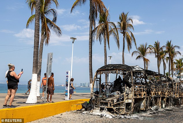 Tourists walk past the burnt wreckage of buses on Monday after a series of blockades and attacks by the cartel following a military operation in which Nemesio Oseguera was killed