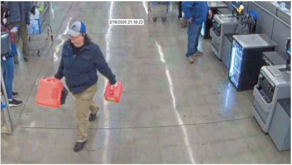 A person carrying two red fuel containers walks through a retail store aisle while customers and checkout machines are visible in the background.