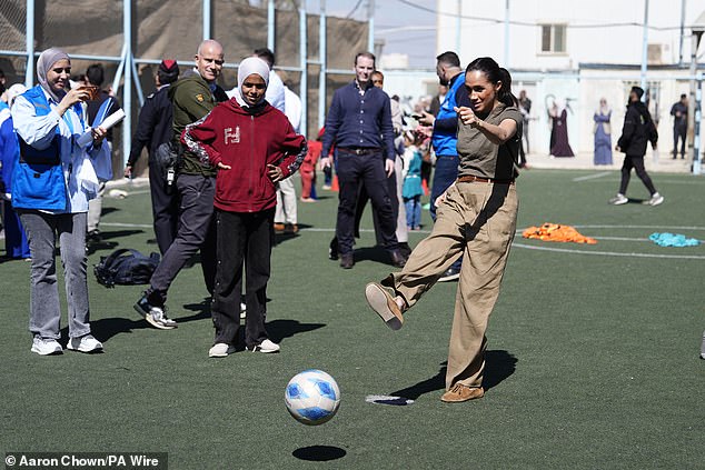 The Duchess of Sussex kicks a ball as a group watch during the tour of Jordan today - an action which body language expert Judi James says was an attempt 'to appear immersive'